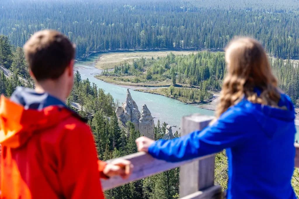 Two hikers in colorful jackets overlook a river winding through lush green forests, with unique rock formations in the distance.